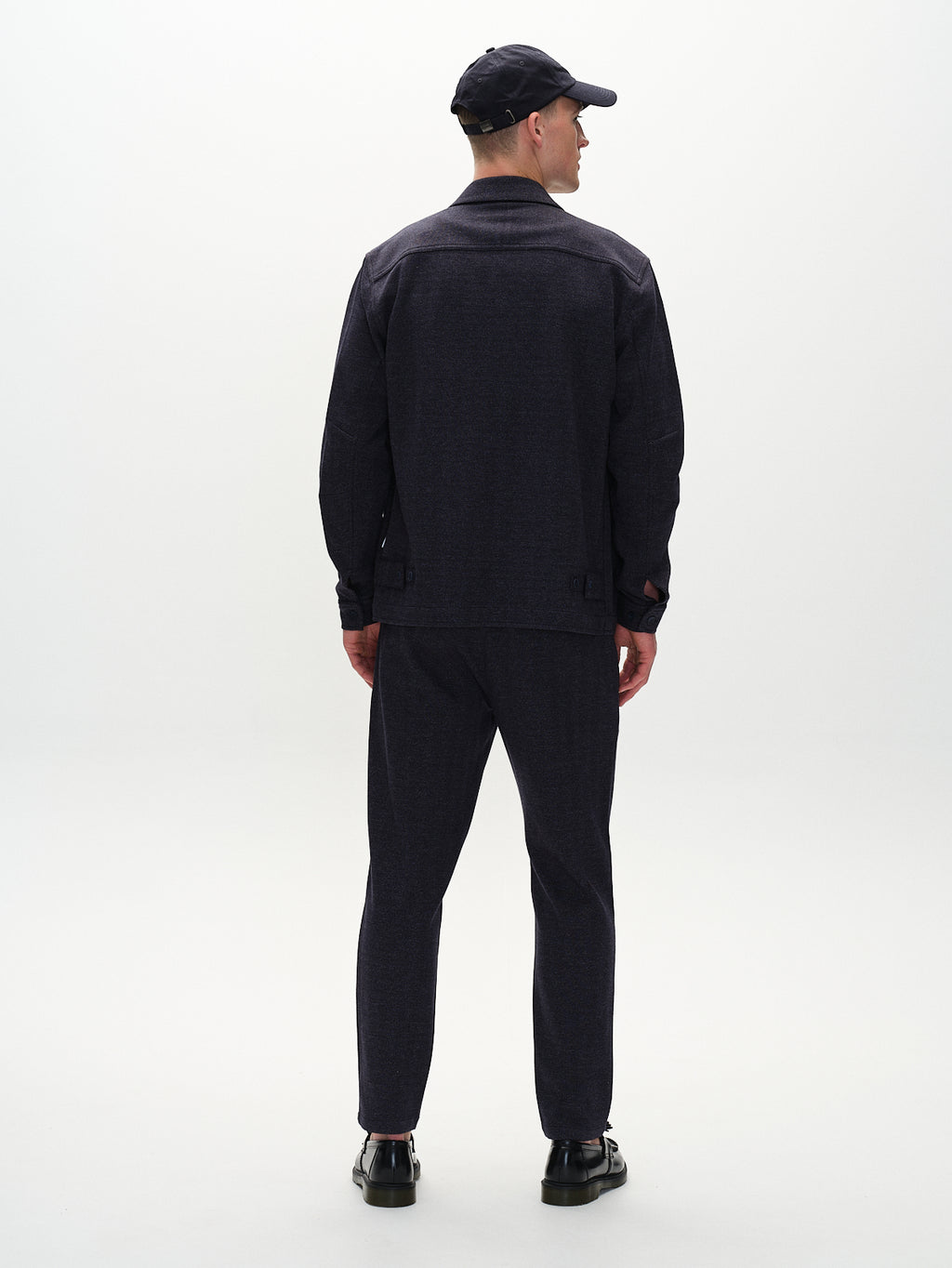 A person in an elegant tweed jacket and Gabbas Monza Tweed - Navy pants stands facing away from the camera against a light gray background, with hands relaxed by their sides. The sophisticated look is completed with dark shoes and a matching cap.