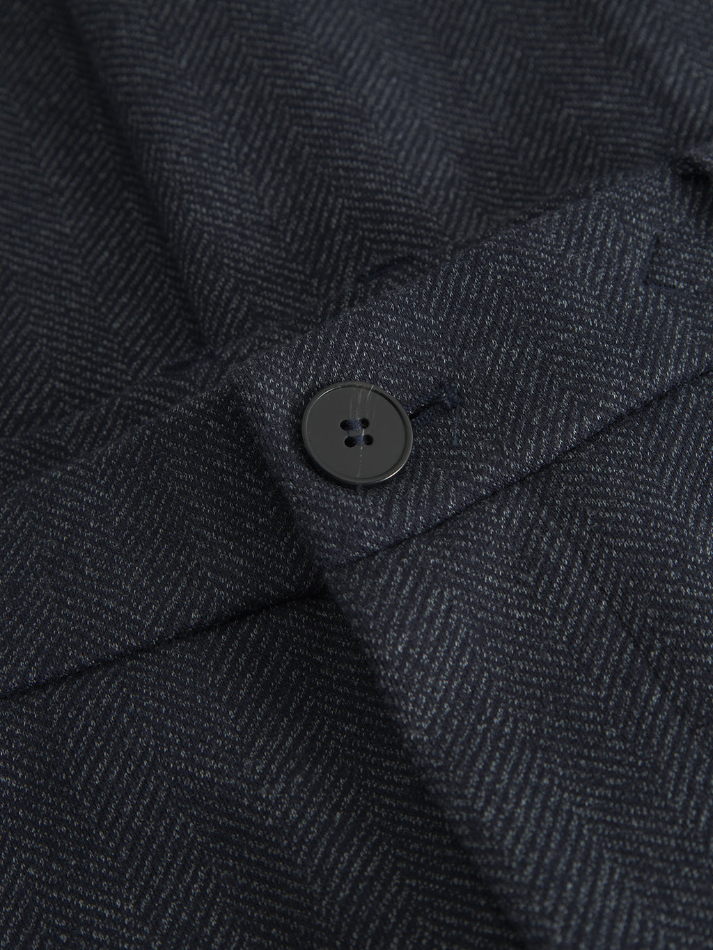 Close-up of Gabbas Monza Tweed - Navy fabric in a dark gray herringbone pattern, highlighting a black button sewn onto it. The diagonal zigzag texture elevates its elegance, with the buttonhole to the right. Ideal for Monza Tweed pants.