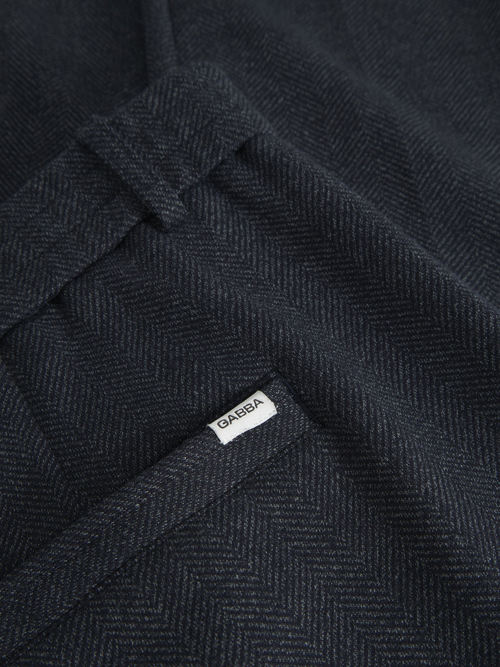 Close-up of dark herringbone fabric with a subtle zigzag pattern, resembling elegant tweed. A small white label reading Gabba is attached near an edge, offering a sophisticated look similar to Monza Tweed - Navy pants.