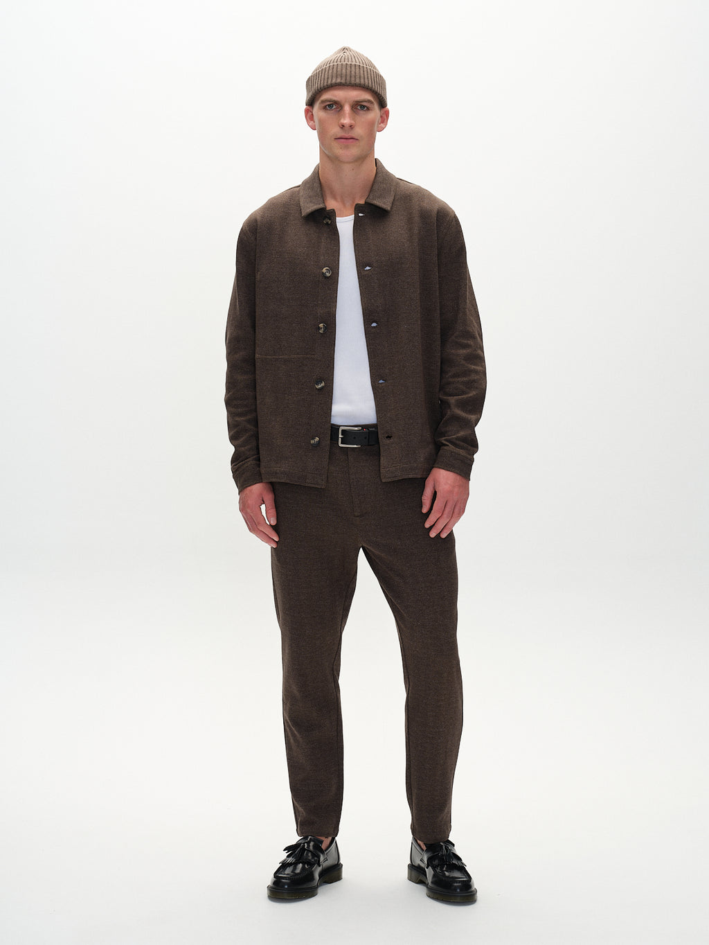 Against a white backdrop, a person models in an elegant brown Monza Tweed button-up shirt by Gabba matched with Monza Tweed pants. A beanie adorns their head, and they layer with a white shirt underneath, paired with black shoes.
