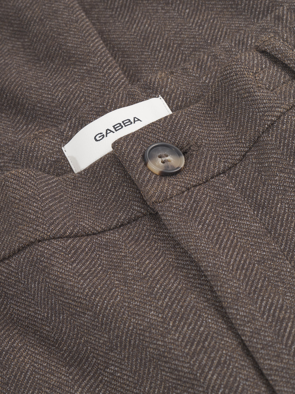 Close-up of a pair of Monza Tweed - Brown pants with a herringbone pattern and button closure. A white Gabba tag is visible above the waistband, enhancing its sophisticated look.