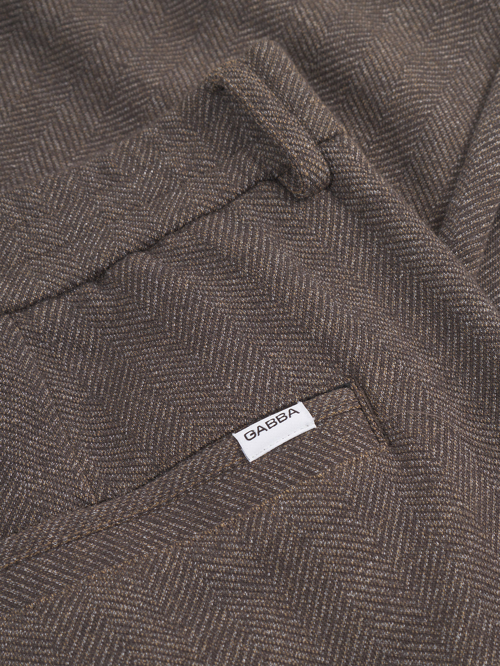 A close-up of brown herringbone fabric reveals a white label with the text Gabba. The detailed tweed texture exhibits a sophisticated pattern in varying shades of brown, reminiscent of Monza Tweed - Brown pants.
