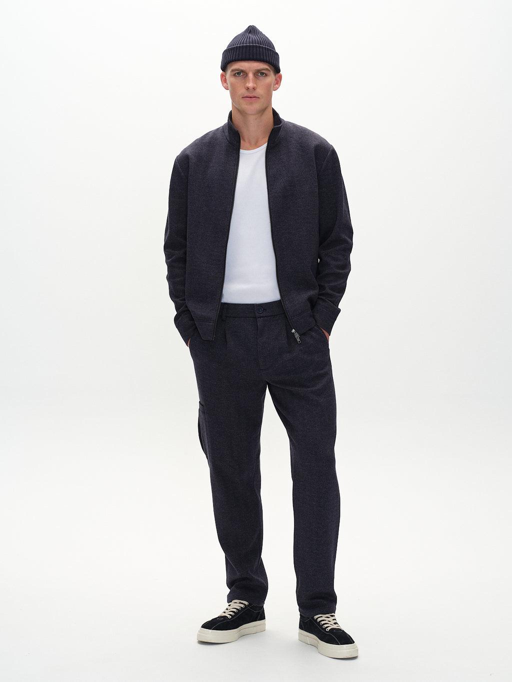 Dressed in Gabbas classic Napoli 19 Tweed - Navy bomber jacket, a white shirt, and matching pants, the person pairs black sneakers with white soles and a cozy knitted beanie while posing against a plain white backdrop.