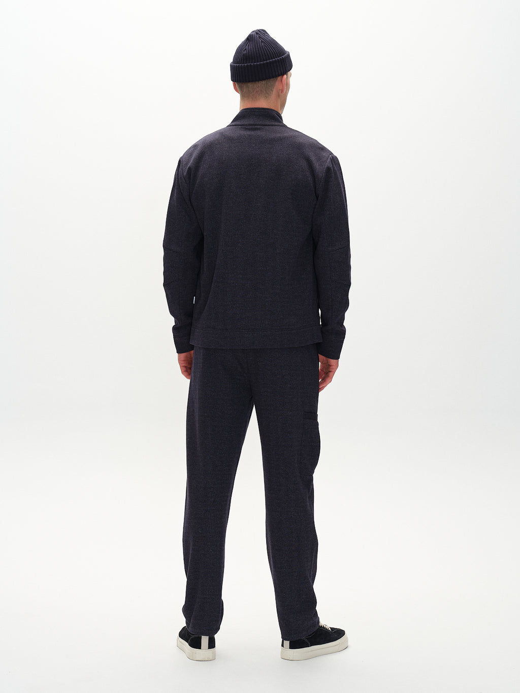 A person wearing Gabbas Napoli 19 Tweed - Navy pants and matching jacket stands with their back to the camera. They sport a beanie and black shoes with white soles, embodying classic style against a plain white background.