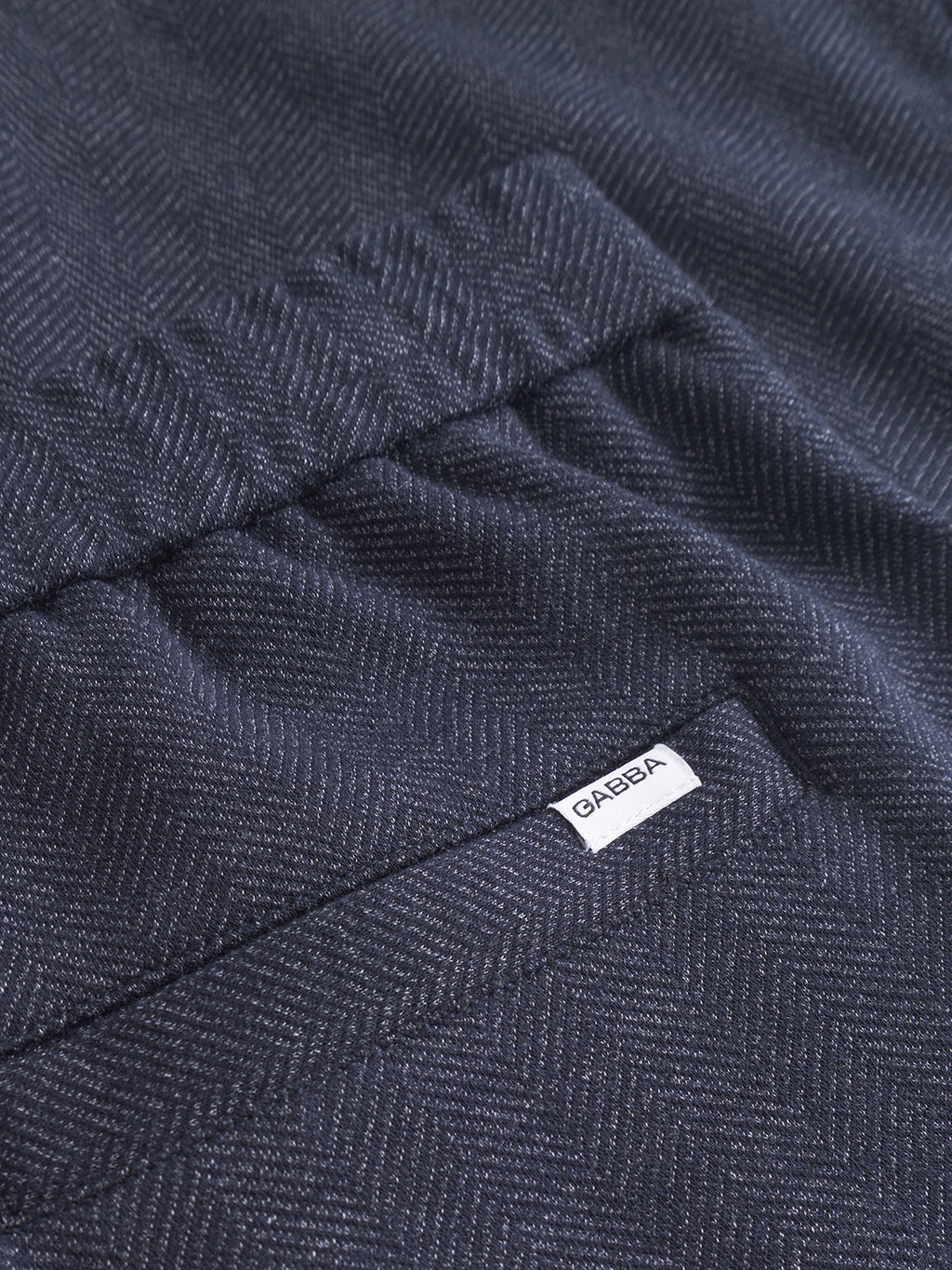Close-up of the classic dark herringbone fabric from the Napoli 19 Tweed - Navy by Gabba, featuring a prominent texture akin to tweed pants. A small white label with GABBA in black text is sewn in the top right corner.