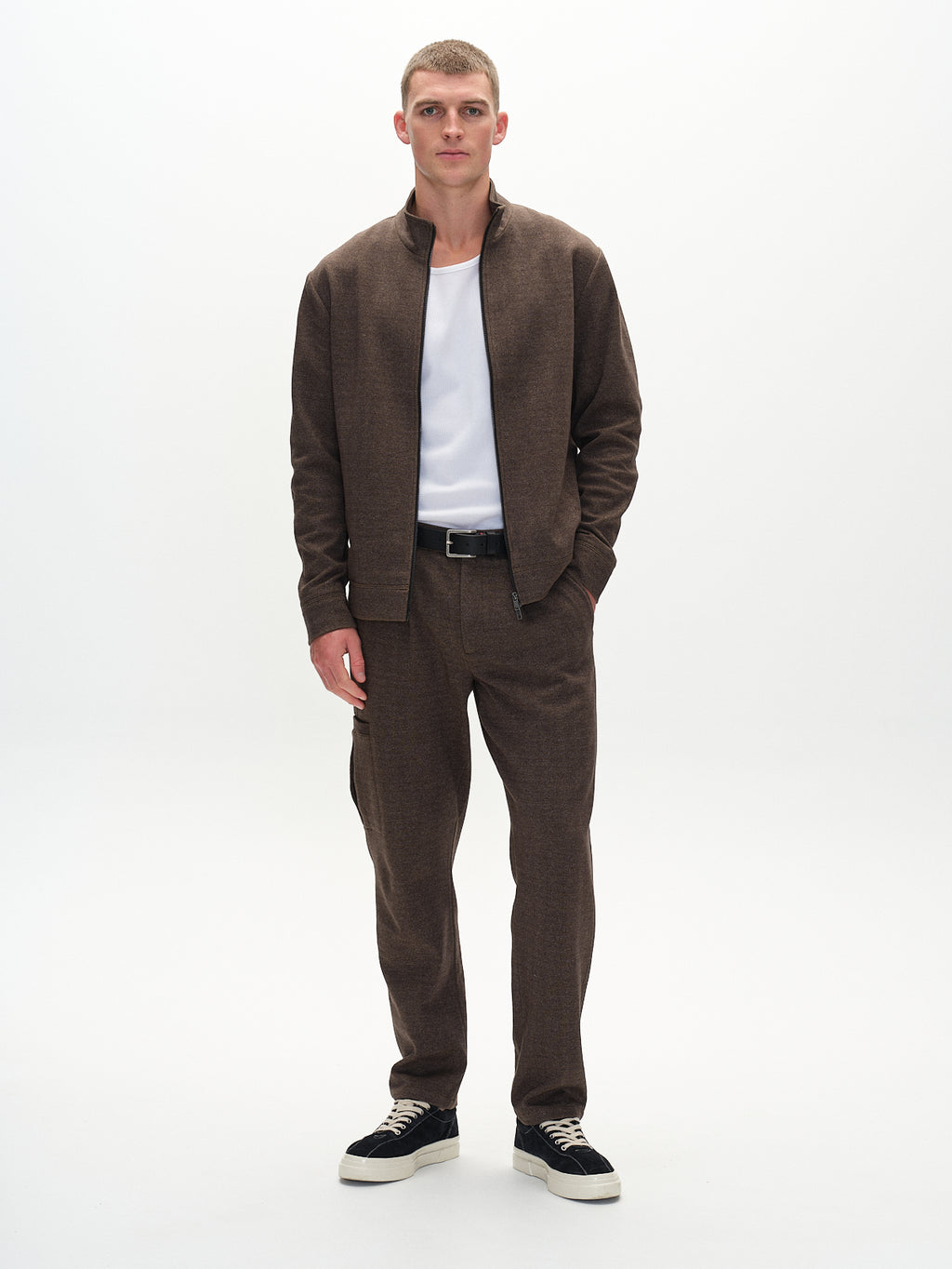 In a light-colored room, a man exudes classic style wearing Gabbas Napoli 19 Tweed trousers and jacket in brown, paired with a crisp white shirt. One hand rests in his pocket, completing the look with black sneakers featuring white soles.