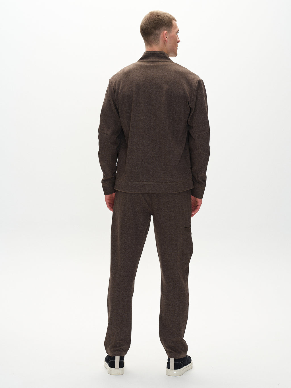 A person stands with their back to the camera, showcasing the classic style of Gabbas Napoli 19 Tweed - Brown jacket and pants. Their short hair is subtly lit in a neutral space, and black-and-white sneakers complete the timeless fashion look.