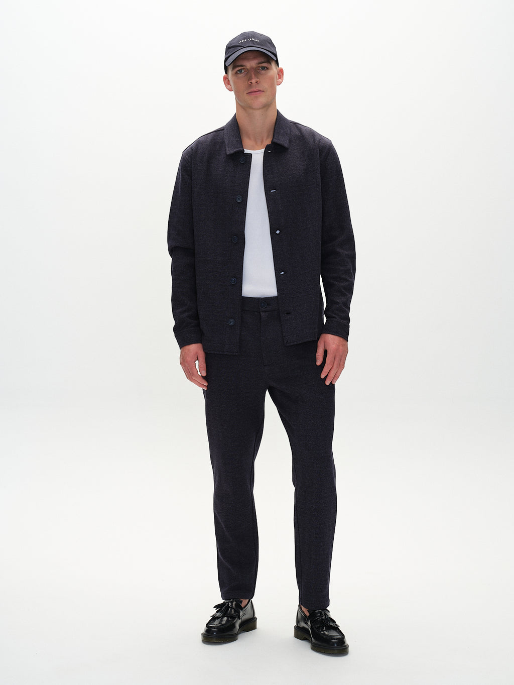A man exudes timeless style against a white background, wearing Gabbas Peter Tweed - Navy jacket and pants, complemented by a crisp white shirt, black loafers, and a black cap. His hands rest casually by his sides.