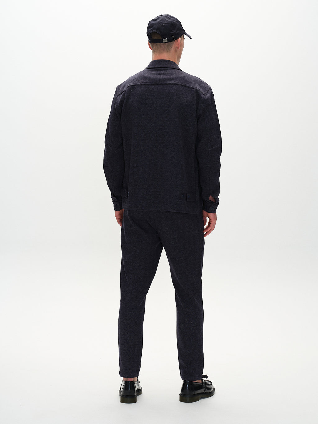 A person wearing Gabba’s Peter Tweed - Navy, including a tailored jacket, matching pants, and shoes stands with their back to the camera in a minimalist setting. Their black cap complements the ensemble against a plain, light background.