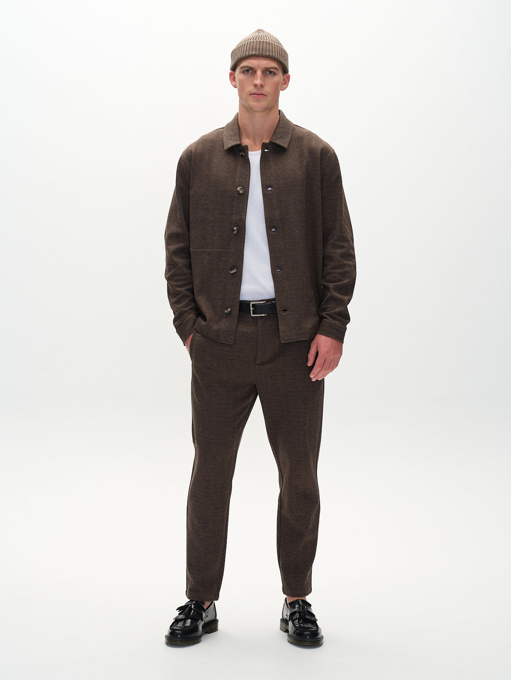 A person poses against a plain backdrop in Gabbas Peter Tweed - Brown jacket and pants, paired with a white T-shirt, beige beanie, and black shoes. With hands in pockets and a neutral expression, they exude sophisticated style.