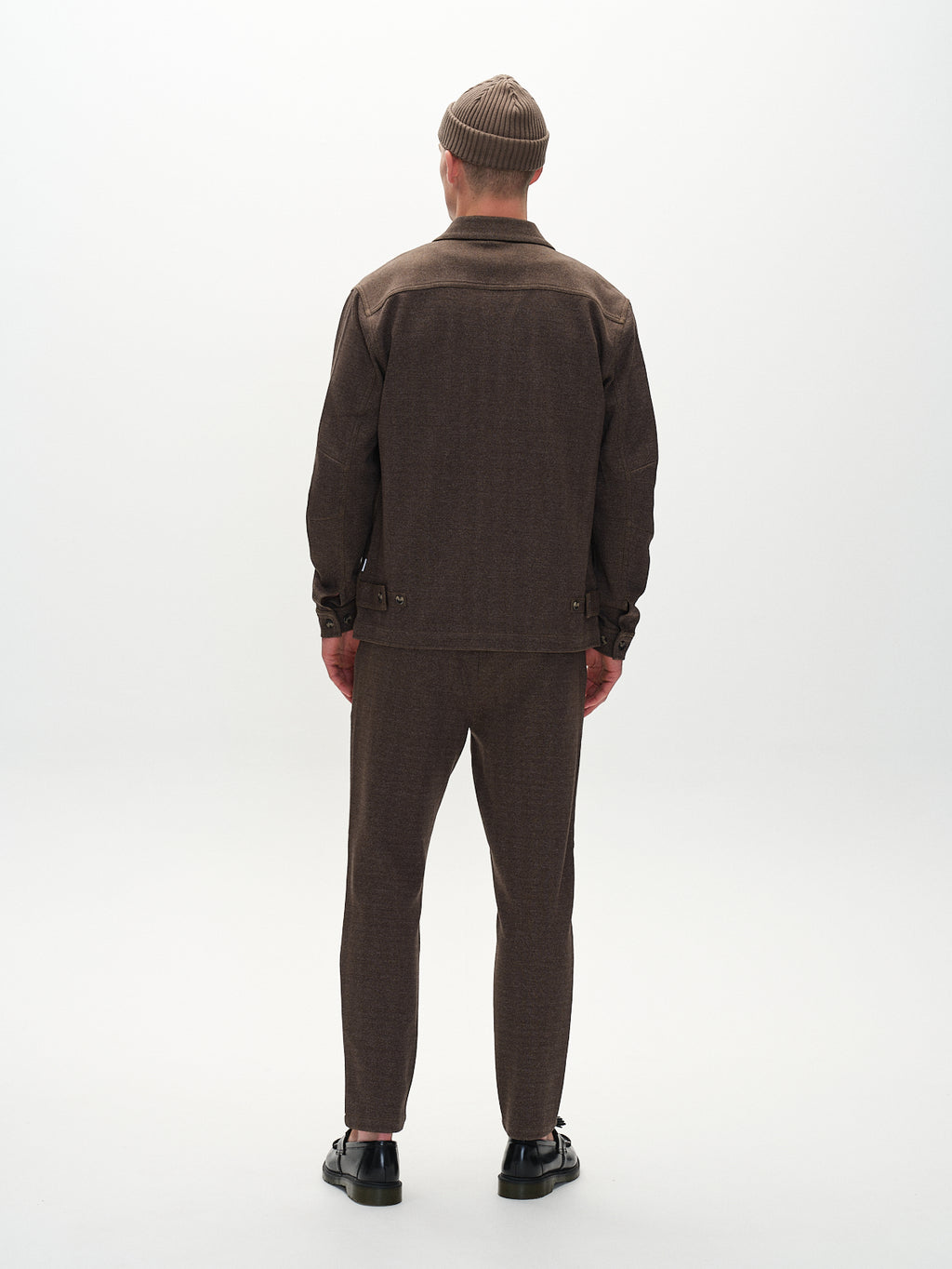 Facing away, a person models the Gabba Peter Tweed - Brown outfit featuring a sophisticated tweed jacket with matching brown pants and beanie. They wear black shoes against a simple, light backdrop.