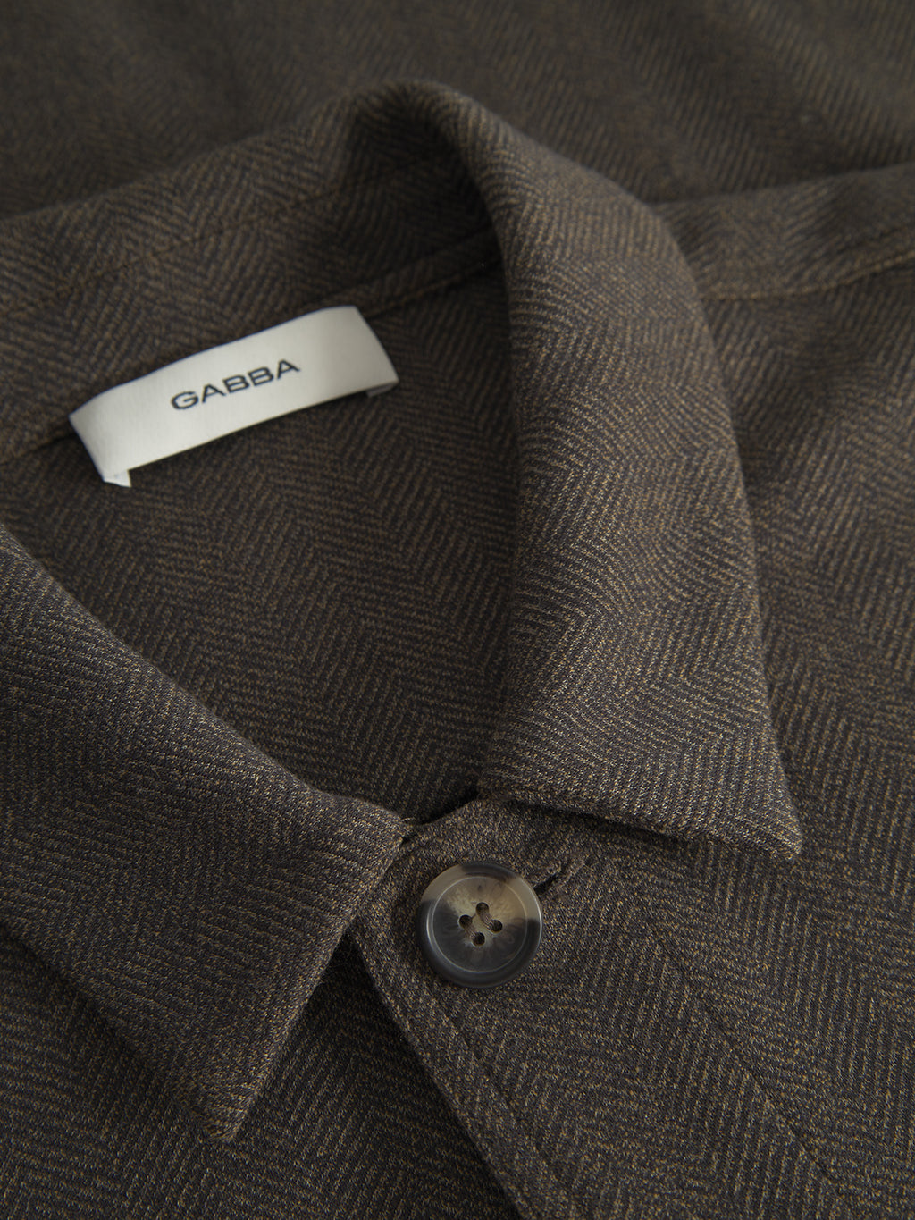 A close-up of the Peter Tweed - Brown jacket by Gabba shows a brown herringbone pattern with a tailored fit, folded collar, and a distinctive white Gabba label. A black button near the bottom edge accentuates its sophisticated design.