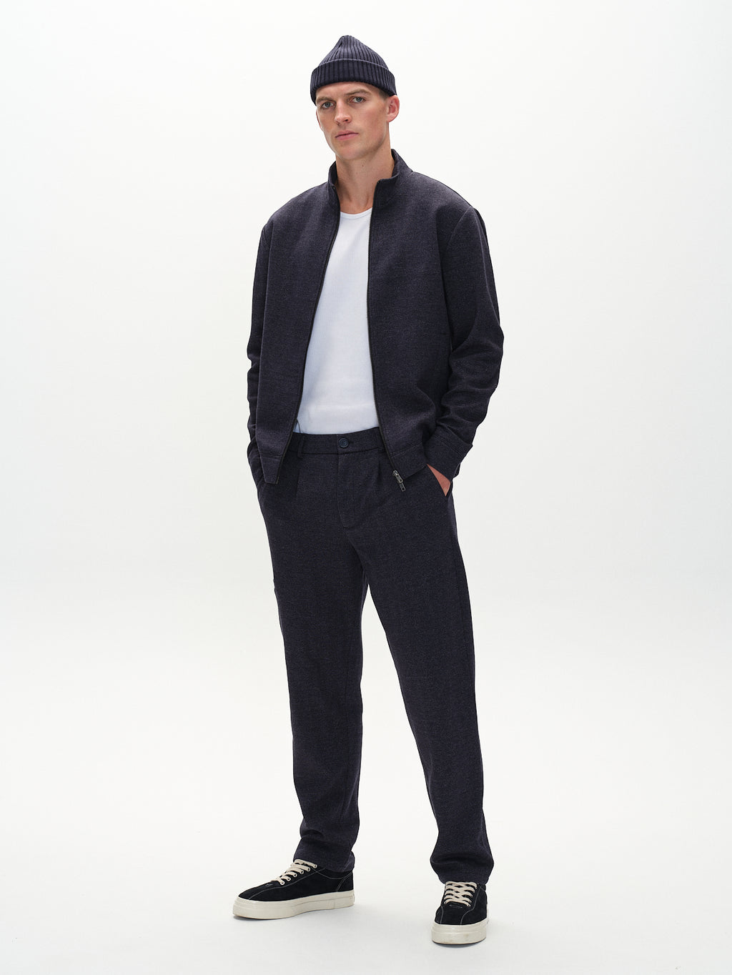 In a well-lit, minimalist space, a person wears the Gabba Nik Tweed - Navy jacket and matching trousers. The rich tweed complements the white shirt and beanie. Black sneakers with white soles complete the look, hands casually tucked into elegant hidden side pockets.