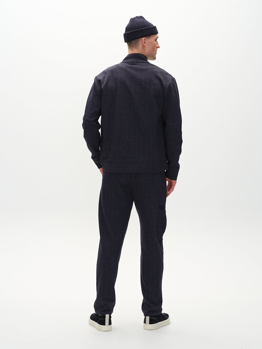 Wearing the Gabba Nik Tweed - Navy jacket with elegant hidden side pockets, a person stands facing away on a light background. They sport a perfectly matching dark beanie, pants, and shoes.