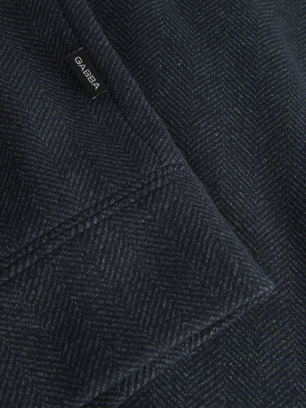 Close-up of a navy herringbone-patterned fabric with a small black tag reading Gabba in white text, showcasing the rich tweed texture of the Nik Tweed jacket by Gabba.