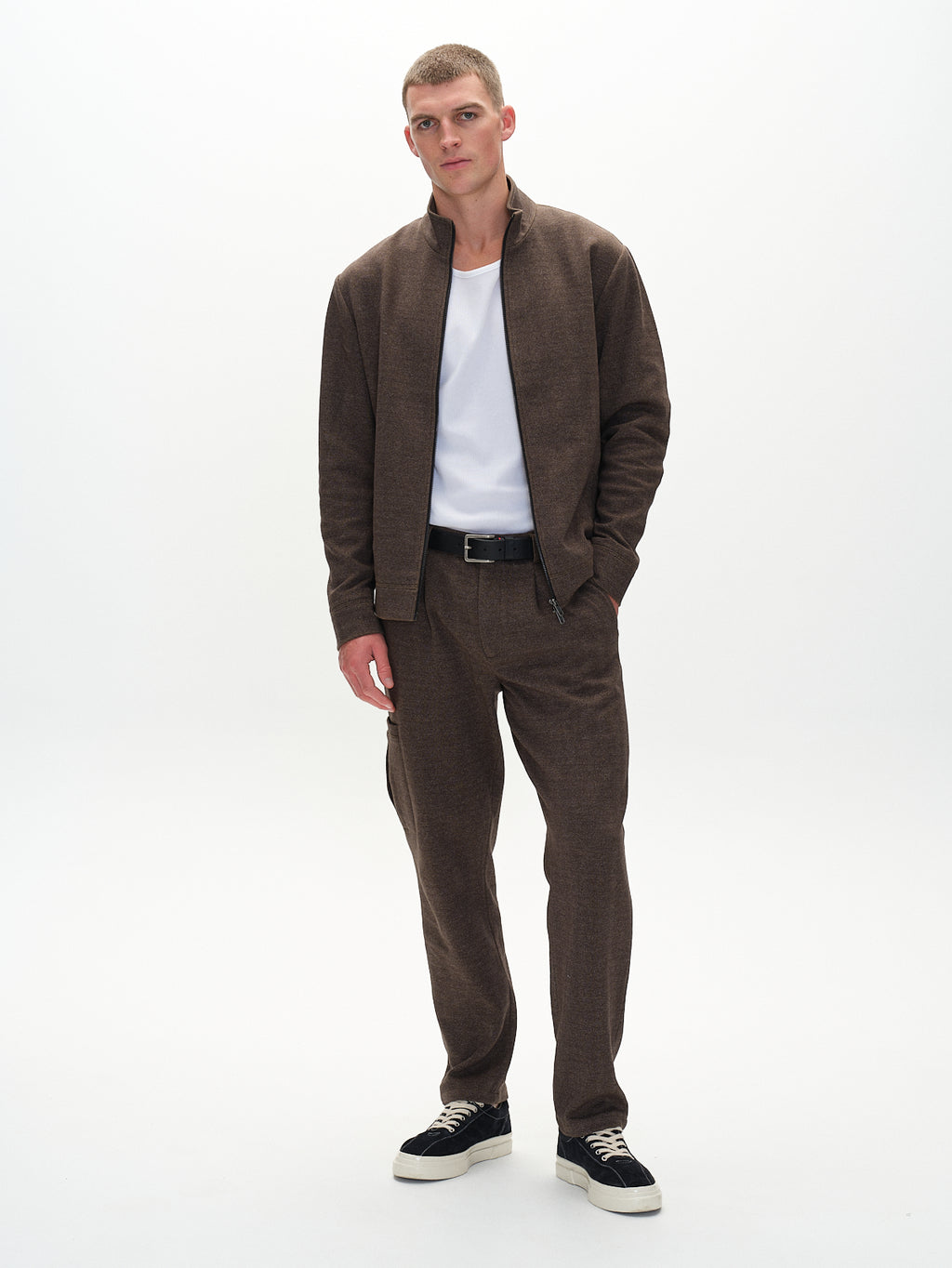 A short-haired individual sports an everyday look in a Gabba Nik Tweed - Brown zip-up jacket over a white shirt, paired with matching pants and black sneakers, posing against a plain white background with one hand in their pocket and the other by their side.