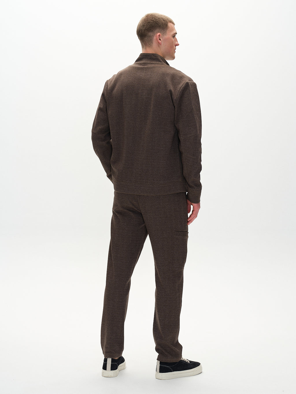 A person models an effortlessly chic look in the Gabba Nik Tweed - Brown jacket and pants, hands in pockets. Black sneakers with white soles complete the outfit, set against a plain white background.