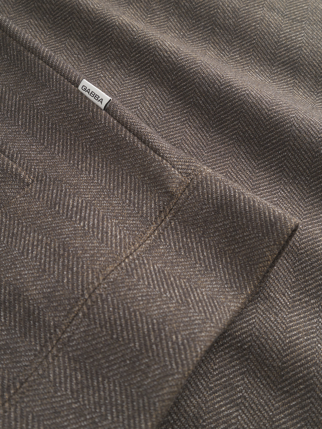 Close-up of Nik Tweed - Brown fabric, featuring a subtle herringbone pattern ideal for everyday wear. A white label with black Gabba text is sewn near the seam, showcasing its texture and stitching.