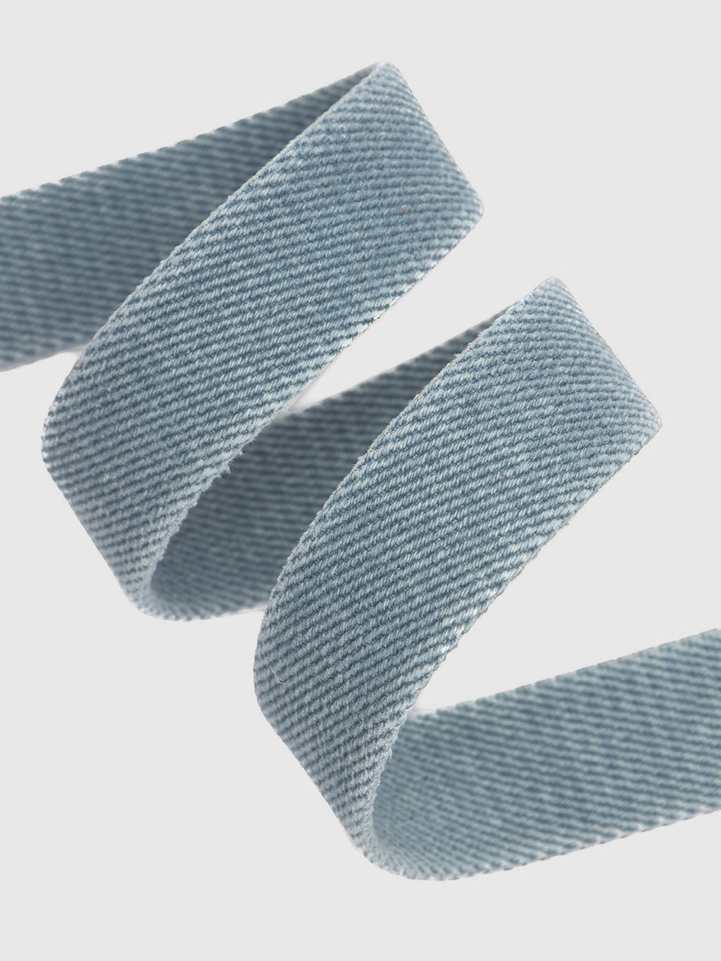 A close-up view of the Gabba Manica Denim Belt in light blue shows its denim-like ribbon curled against a white background, revealing a diagonal weave pattern reminiscent of high-quality denim fabric.