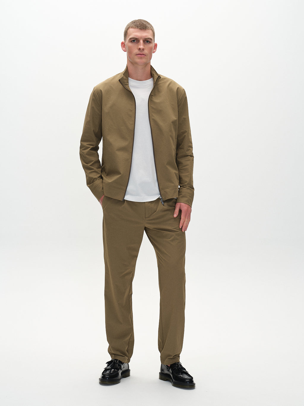 Wearing a light brown jacket and Gabbas Napoli 19 Tech - Army pants over a white T-shirt, a person with short hair stands comfortably. Their hands are in their pockets, complemented by black shoes against a plain white background as they gaze slightly to the side.