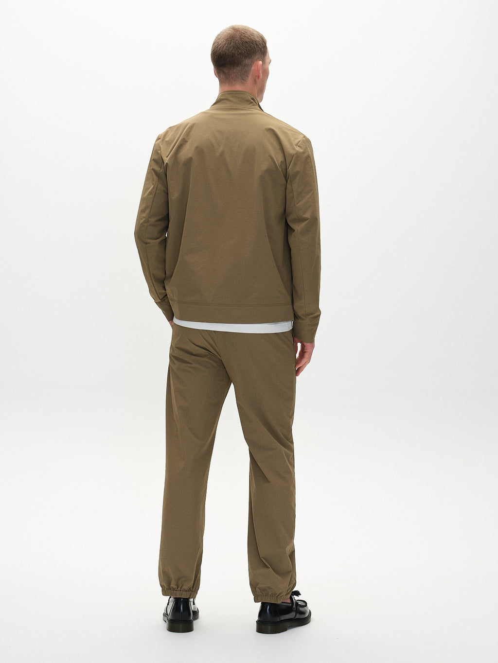 A person faces away from the camera, wearing a light brown jacket and Gabbas Napoli 19 Tech - Army pants for a versatile look. A white shirt peeks out underneath and is paired with black shoes, all set against a plain white background to highlight comfort and minimalism.