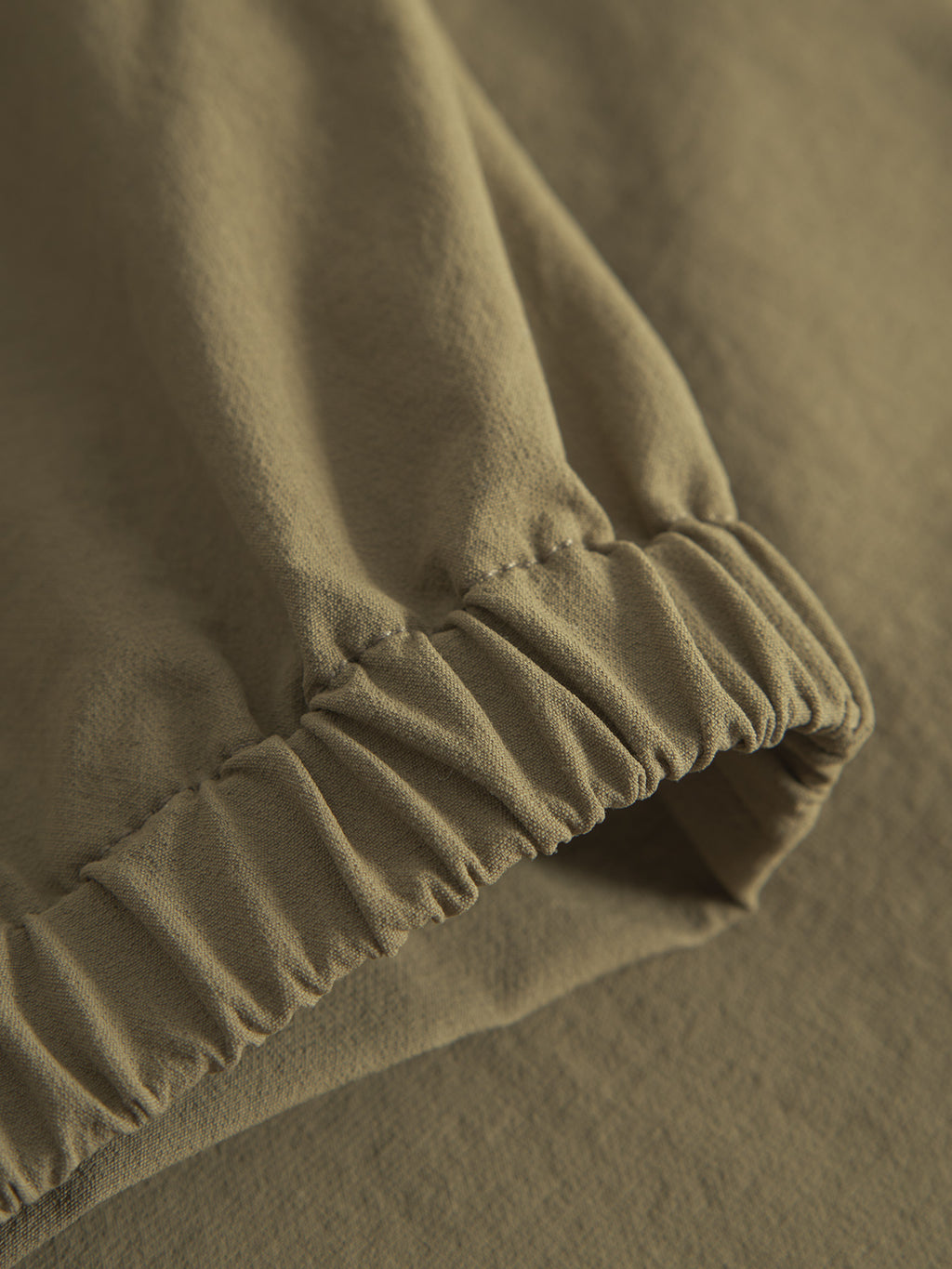 A close-up of the Napoli 19 Tech - Army fabric by Gabba reveals a soft, slightly textured khaki green material with an elasticated seam, hinting at its versatile design. The natural folds promise comfort.