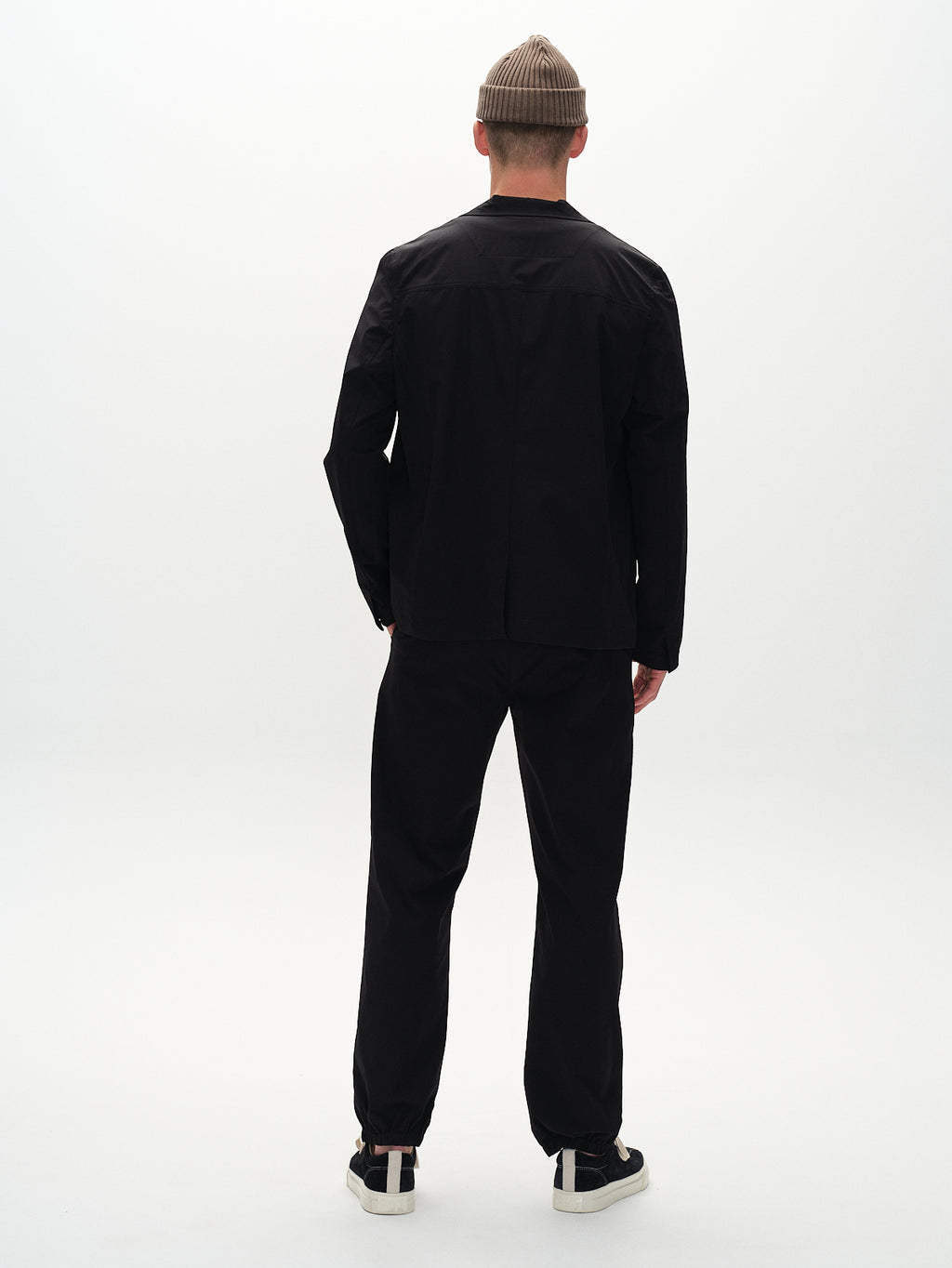 A person stands with their back to the camera, wearing a black suit paired with Gabbas Napoli 19 Tech - Black pants and black sneakers for a versatile design. Topped with a beige beanie, they embody comfort against a plain white backdrop.