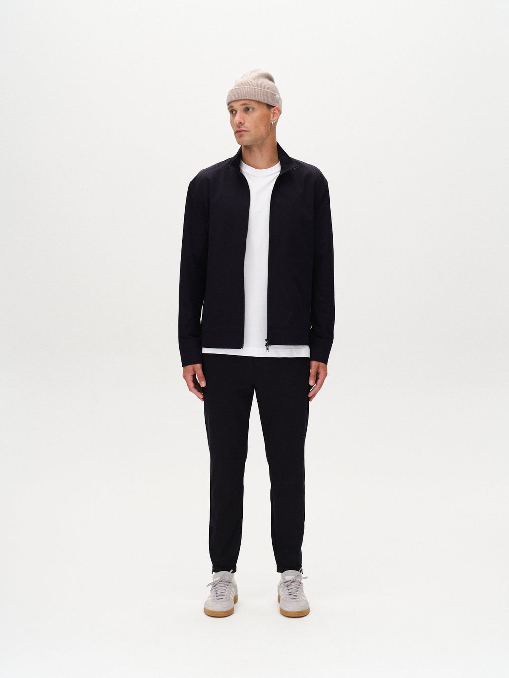 A man stands against a white background wearing Gabbas Nik Tech jacket in navy, a light gray beanie, white t-shirt, black pants, and light sneakers—an effortlessly stylish look for an on-the-go lifestyle. He looks slightly to his left.