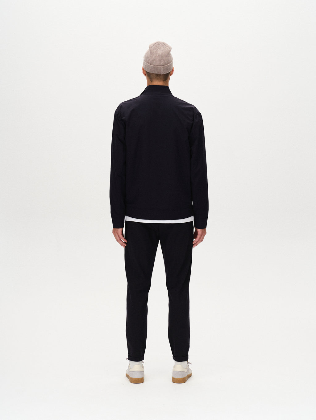 A person wearing a light gray beanie, Gabba Nik Tech navy zip-front jacket, black pants, and white sneakers stands facing away against a plain white background.