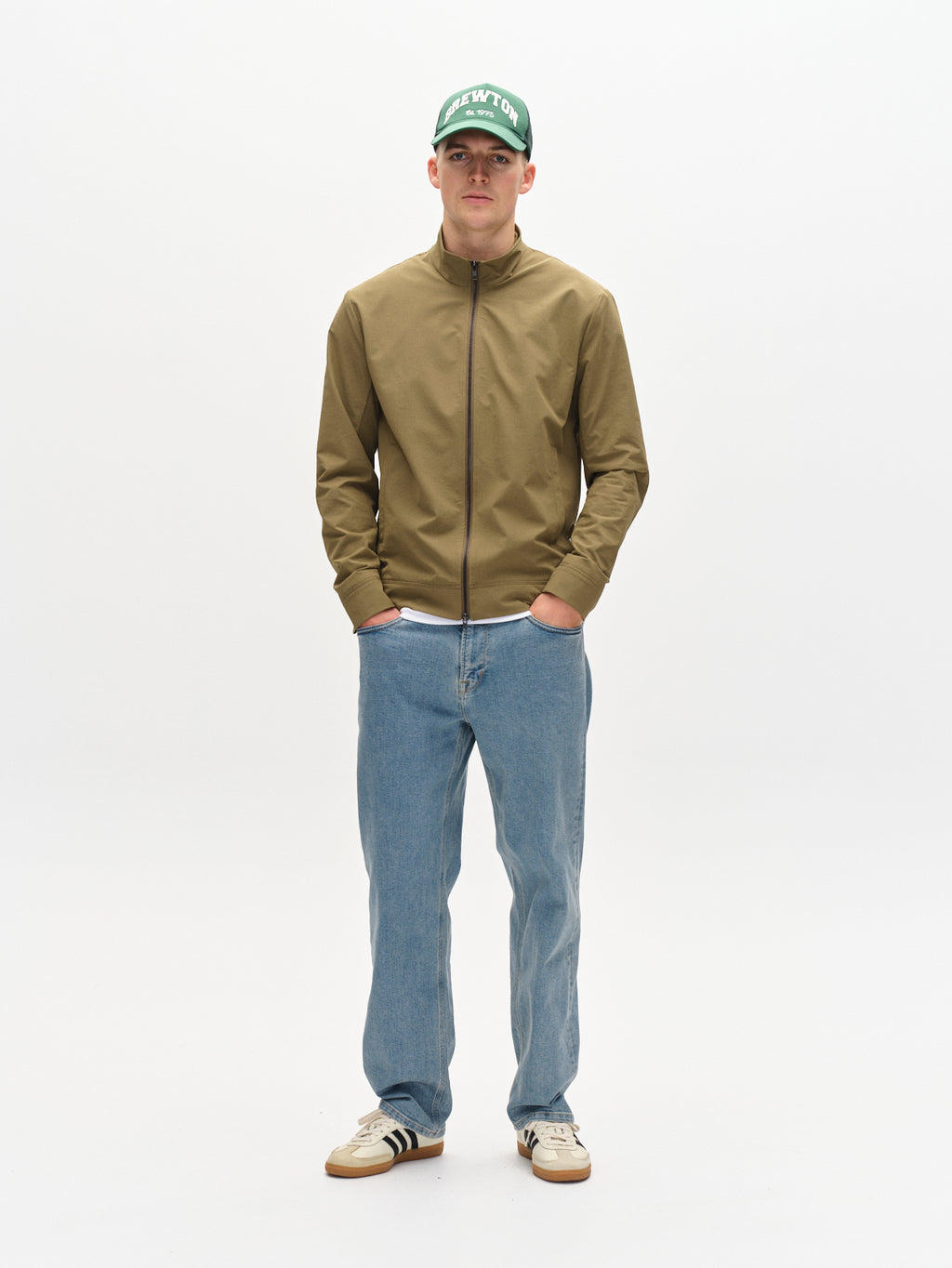 Wearing Gabbas Nik Tech - Army cap, a person stands against a plain white background. They sport a khaki zip-front jacket, blue jeans, and white sneakers with black stripes. Their hands rest in their pockets as they face the camera, embodying an on-the-go lifestyle.