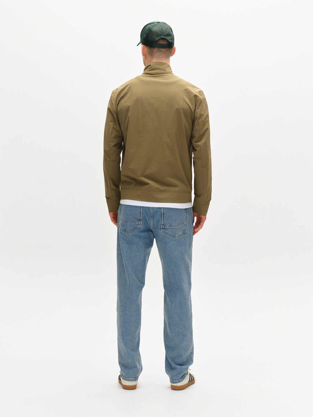 A person exudes an on-the-go lifestyle against a white background, wearing the Nik Tech - Army green cap by Gabba, a brown zip-front jacket, light blue jeans, and striped sneakers.
