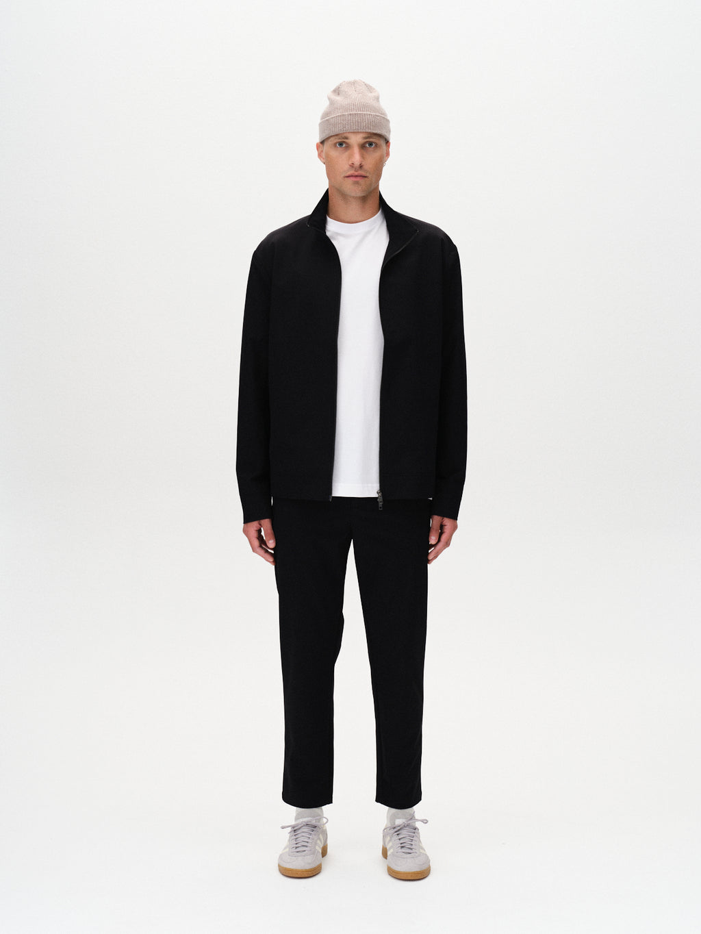 A man stands facing forward against a plain white background wearing a beige beanie, white t-shirt, black pants, white sneakers, and the Gabba Nik Tech - Black jacket with a practical design.