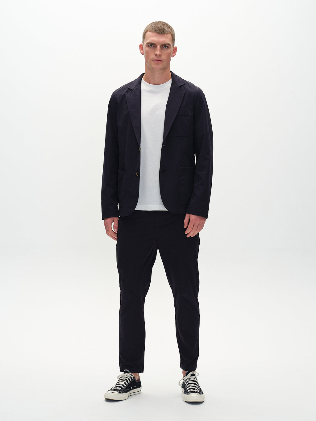 In a minimalist setting, someone wears Gabbas Plaza Tech jacket in navy over a tailored white T-shirt, paired with black pants and black sneakers featuring white soles. This creates a stylish contrast against the plain white background.
