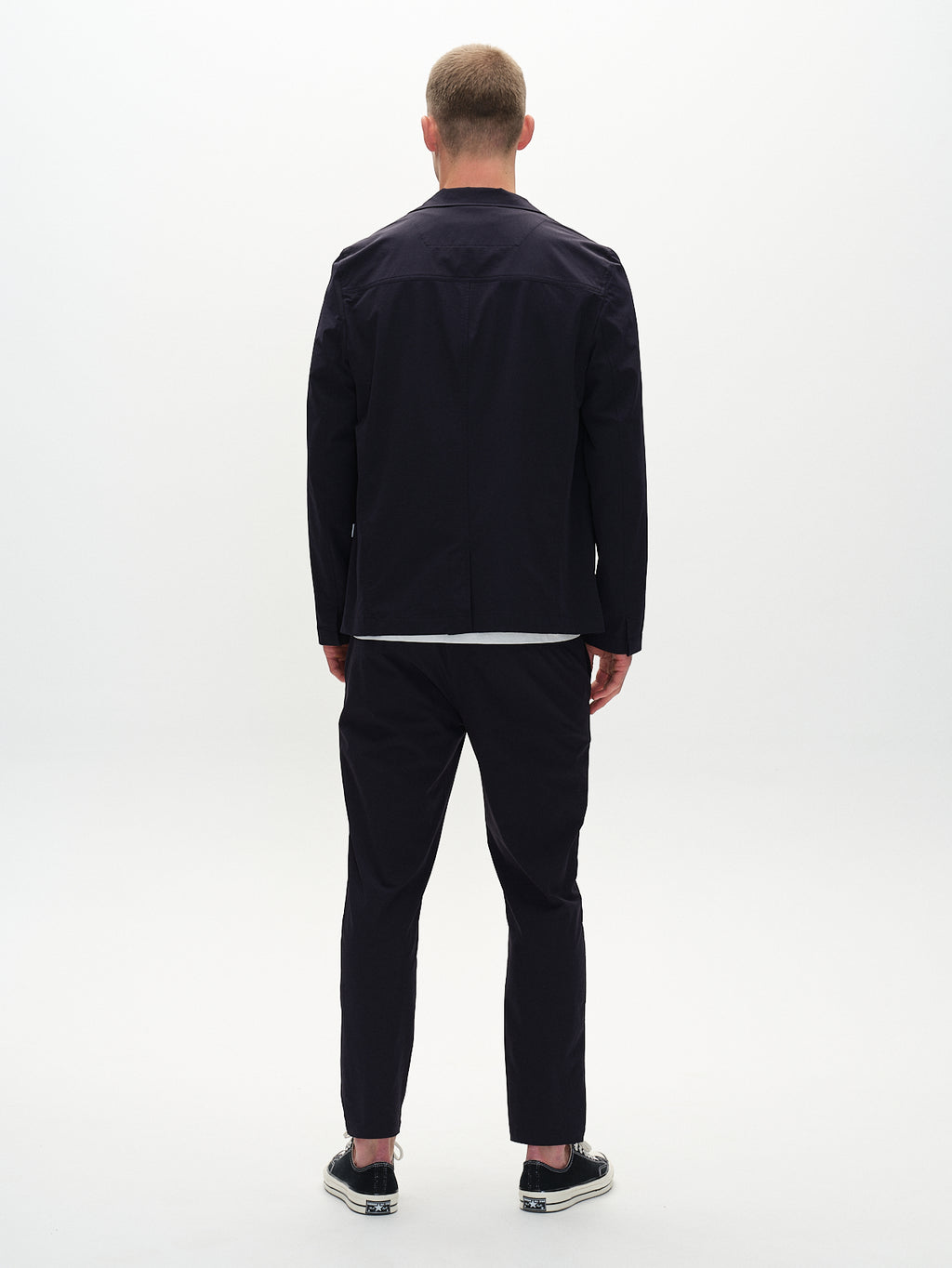 A person stands with their back to the camera in a Gabba Plaza Tech - Navy jacket and pants, tailored to fit. They have short hair and wear black sneakers with white soles against a plain white background.