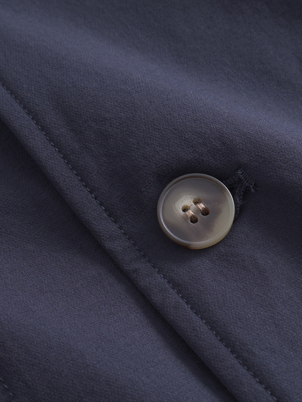 Close-up of the Plaza Tech - Navy fabric from Gabba, featuring a single glossy beige button with four holes against the matte dark blue texture. A diagonal seam runs across this tailored piece, emphasizing its smooth surface.