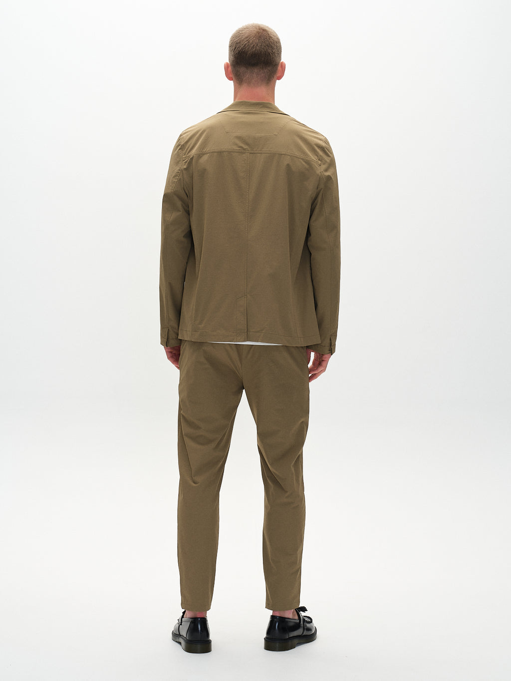 Facing away, a person wears Gabbas Plaza Tech - Army jacket and pants in olive-green with a tailored fit. Against a white background, they pair it with black sandals. The minimalist outfit features clean lines and spacious pockets, blending functionality with style effortlessly.