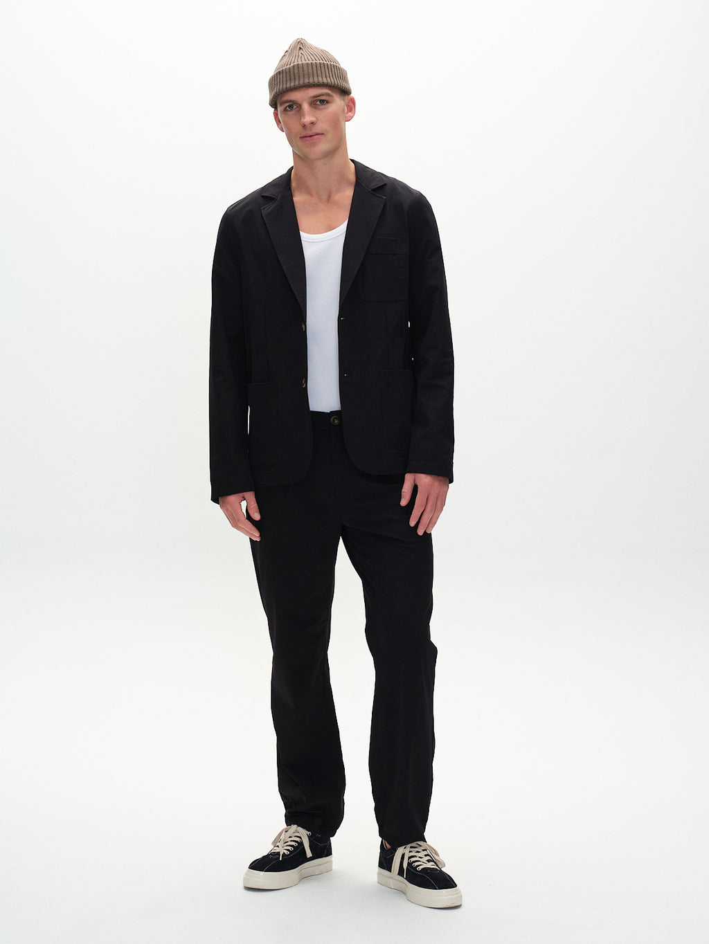A person in Gabbas Plaza Tech - Black tailored fit suit jacket and pants, paired with a white shirt and black sneakers, stands against a plain white background. Theyre wearing a beige knitted hat and look at the camera, perfectly balancing style with functionality.
