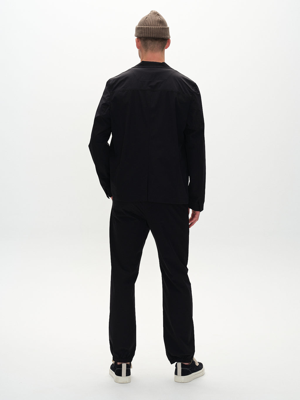 A person wearing Gabbas Plaza Tech black jacket and pants stands with their back to the camera. They sport a beige beanie and black sneakers with white soles, achieving a tailored look that contrasts perfectly against the plain white background.