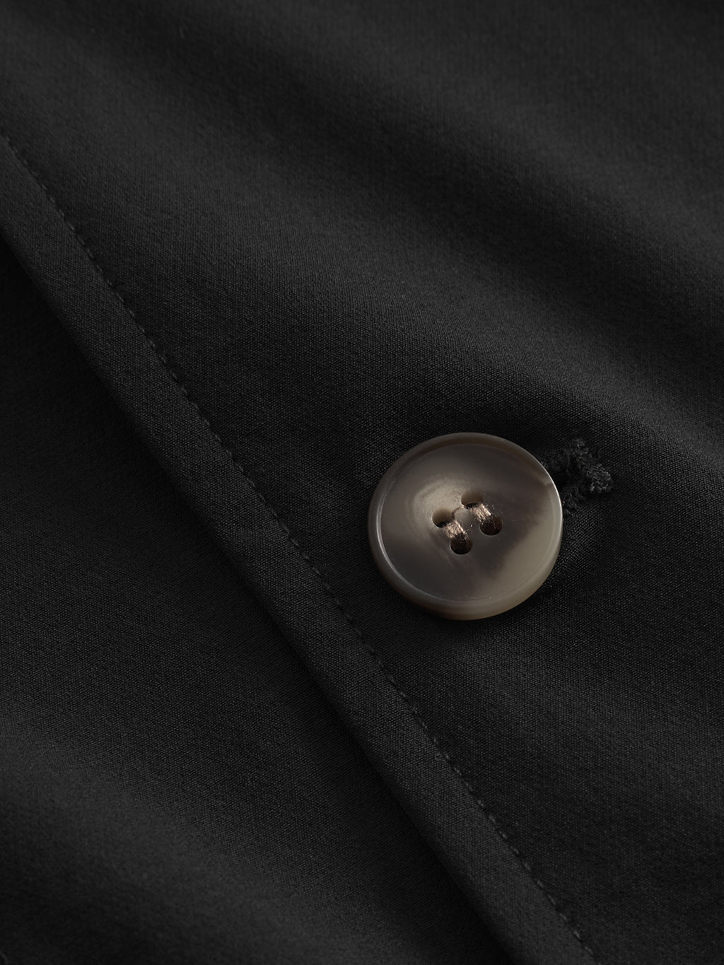 A close-up of the Gabba Plaza Tech - Black jacket highlights its glossy dark gray button sewn onto the slightly shiny fabric. The stitched buttonhole offers an exquisite touch by creating a fold, emphasizing the tailored fit details of this tech jacket.