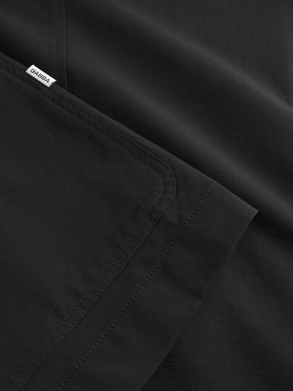 Close-up of the Gabba Plaza Tech - Black jackets neatly folded dark fabric, featuring visible stitching and a small white label. The smooth texture and slightly reflective surface suggest a tailored fit, showcasing its modern design.