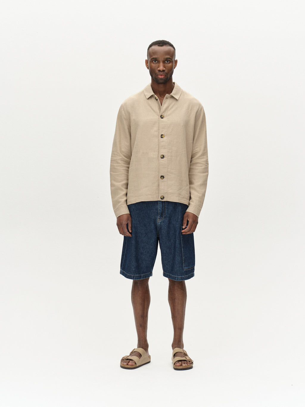 A person stands against a plain background, wearing a beige button-up shirt and Gabbas Ugo Cargo Fone shorts in Mid Blue Denim with a laid-back fit, paired with casual sandals. Their hands rest at their sides, capturing an effortless workwear vibe.