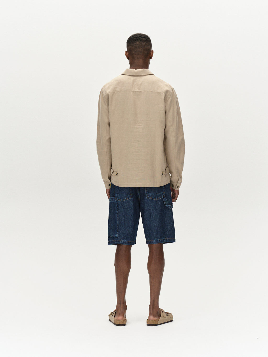 Facing away, someone wears a beige jacket and Gabbas Ugo Cargo Fone - Mid Blue Denim shorts in a loose fit style, paired with sandals. The plain, light-colored background highlights the casually curated outfit.