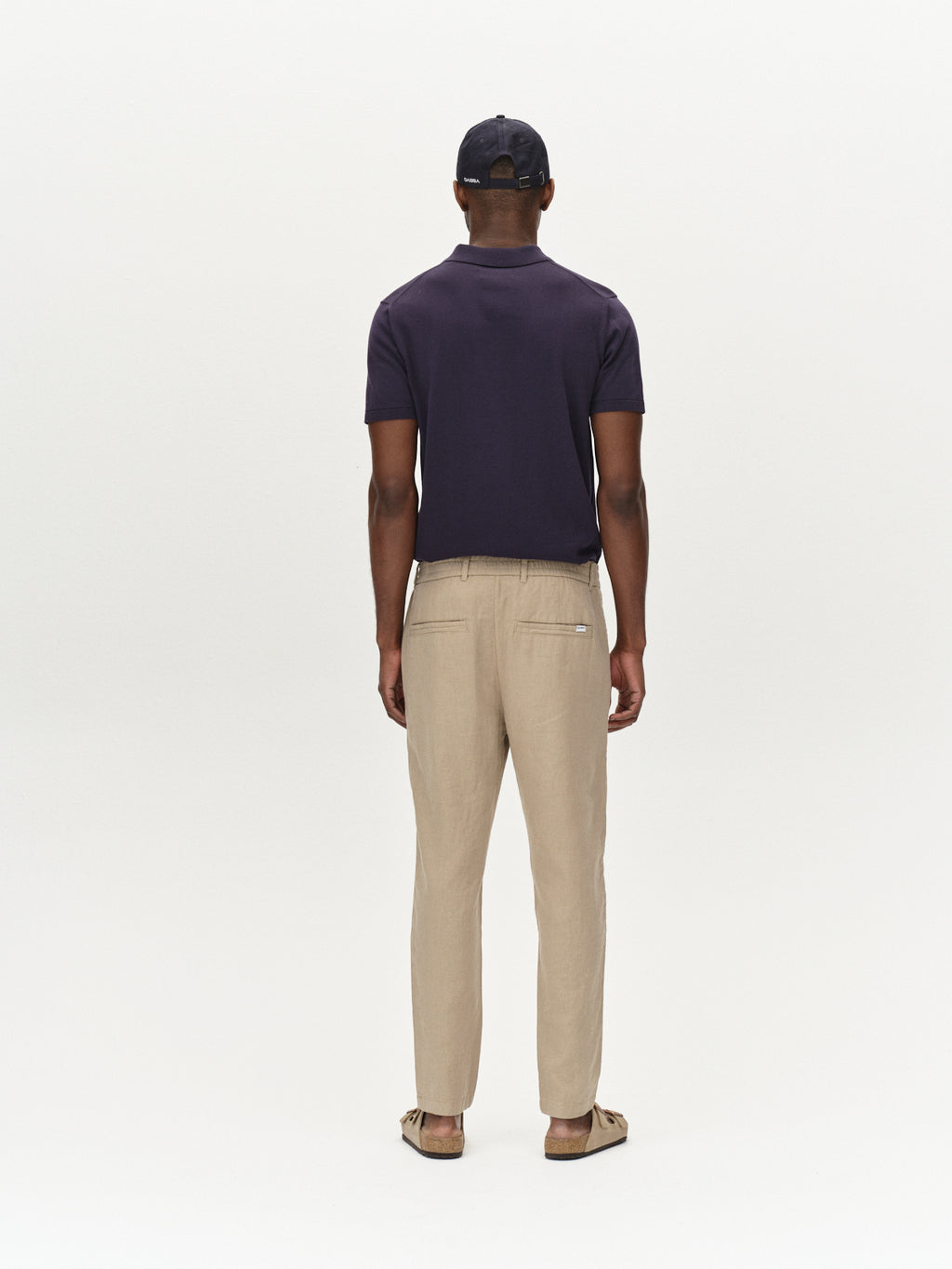 A man faces away from the camera, wearing Gabba’s Monza Lino Cropped - Sand Plaza pants with a navy short-sleeve shirt, tan shoes, and a black cap. The relaxed silhouette and comfort stand out against a plain white background.
