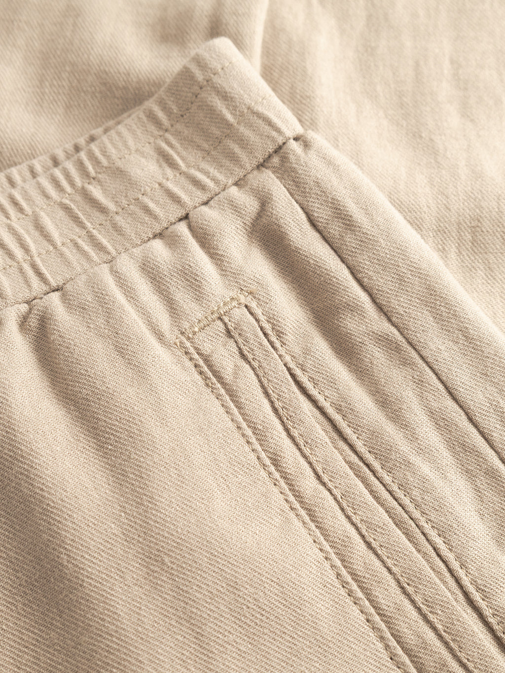Close-up of Gabba Monza Lino Cropped pants in Sand Plaza: beige fabric with a relaxed silhouette, elastic waistband, and a neatly sewn back pocket with a horizontal slit. Texture and stitching showcase comfort and quality craftsmanship.