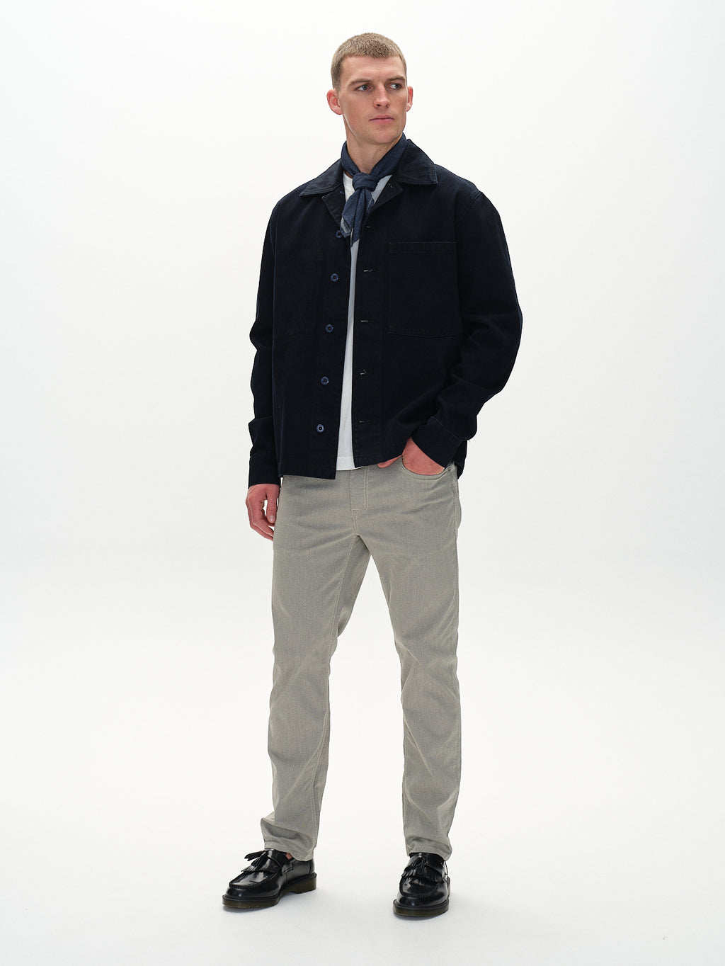 In front of a simple white background, someone is wearing a dark jacket over a gray shirt, Marc K3280 Dale - Birch pants by Gabba in light gray, black shoes, and an elegantly tied blue scarf. As they gaze aside, their regular straight fit ensemble exudes quiet confidence.