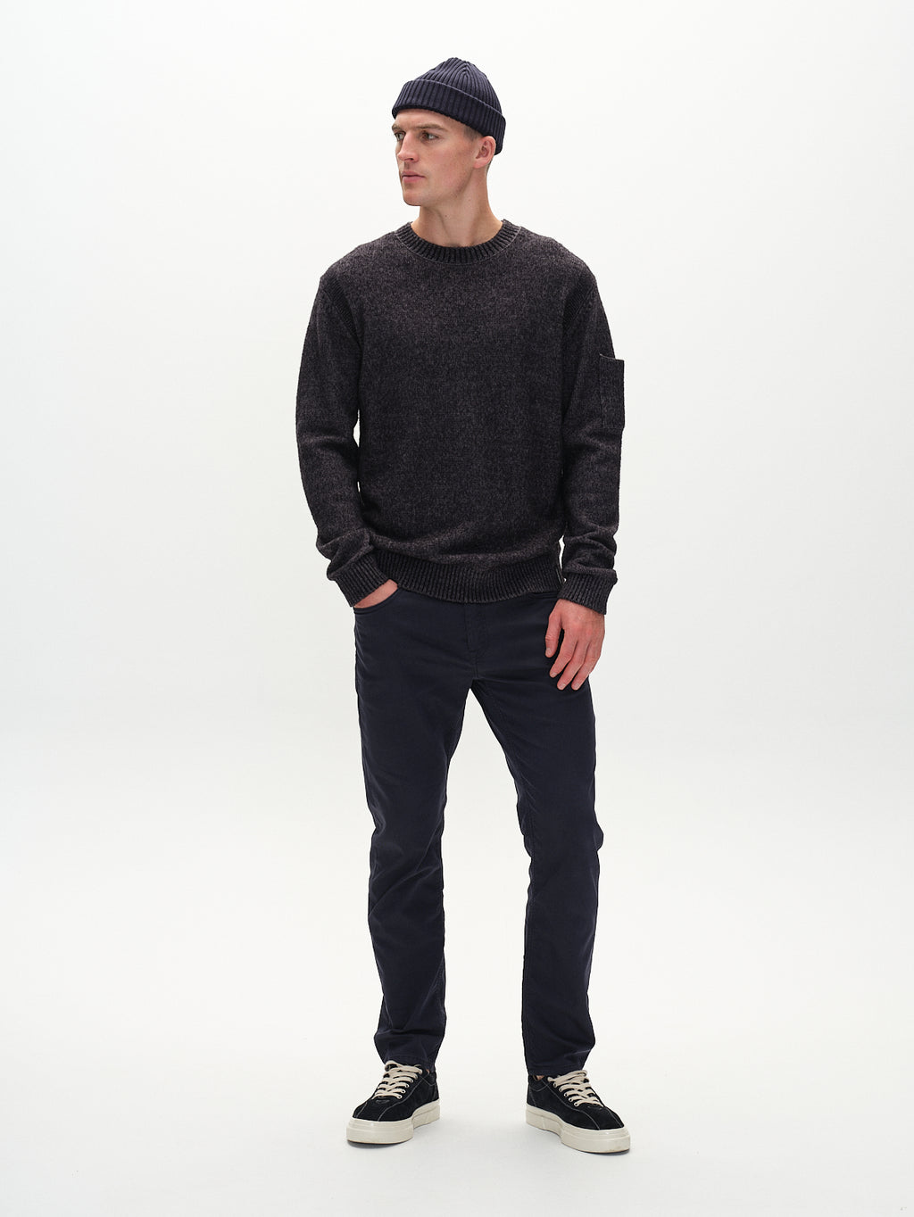 Against a plain white backdrop, an individual dons a dark beanie, charcoal sweater, and Marc K3280 Dale - Navy pants from Gabba. The regular straight fit with Gabba stitching conveys a casual, relaxed style. They pair the outfit with sneakers featuring white soles while looking sideways.