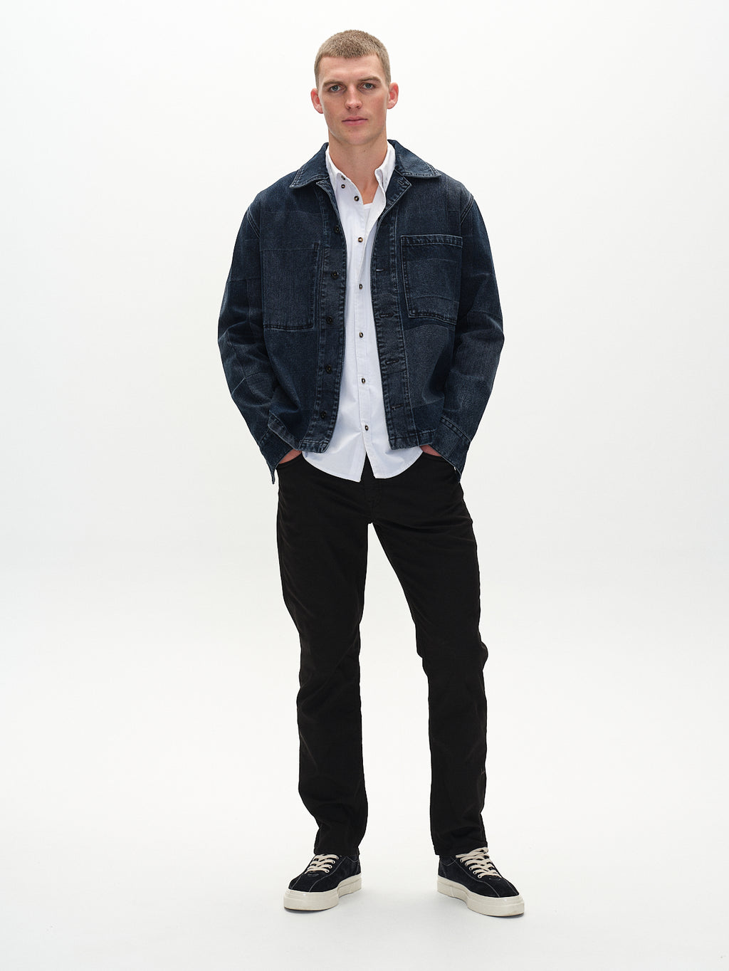 A person stands against a white background, wearing a Gabba dark denim jacket, over a white button-up shirt, paired with regular straight fit black pants. Theyre sporting Marc K3280 Dale - Black sneakers with white soles and appear relaxed with hands in jacket pockets.
