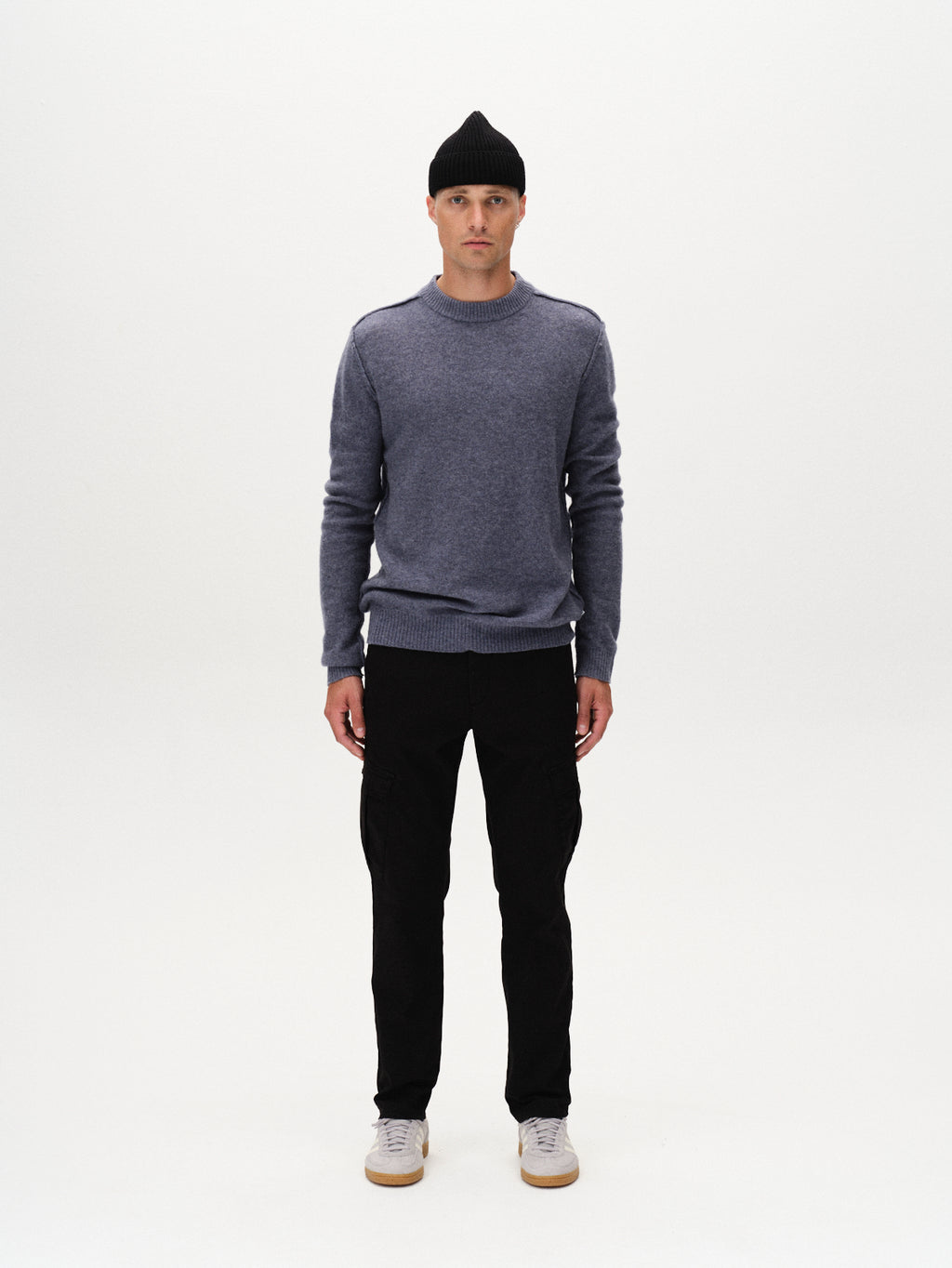 A man stands facing forward against a plain white background, wearing Gabbas Paul K3280 Cargo Dale - Black slim fit cargo pants with functional pockets, paired with a black beanie, blue sweater, and white sneakers. His arms rest at his sides.