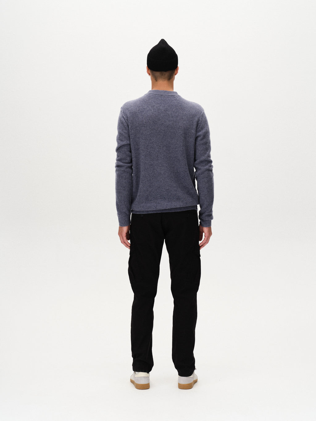 A person stands with their back to the camera, wearing a black beanie, a gray long-sleeve sweater, white shoes, and Gabba Paul K3280 Cargo Dale - Black pants with cargo pockets, against a plain white background.