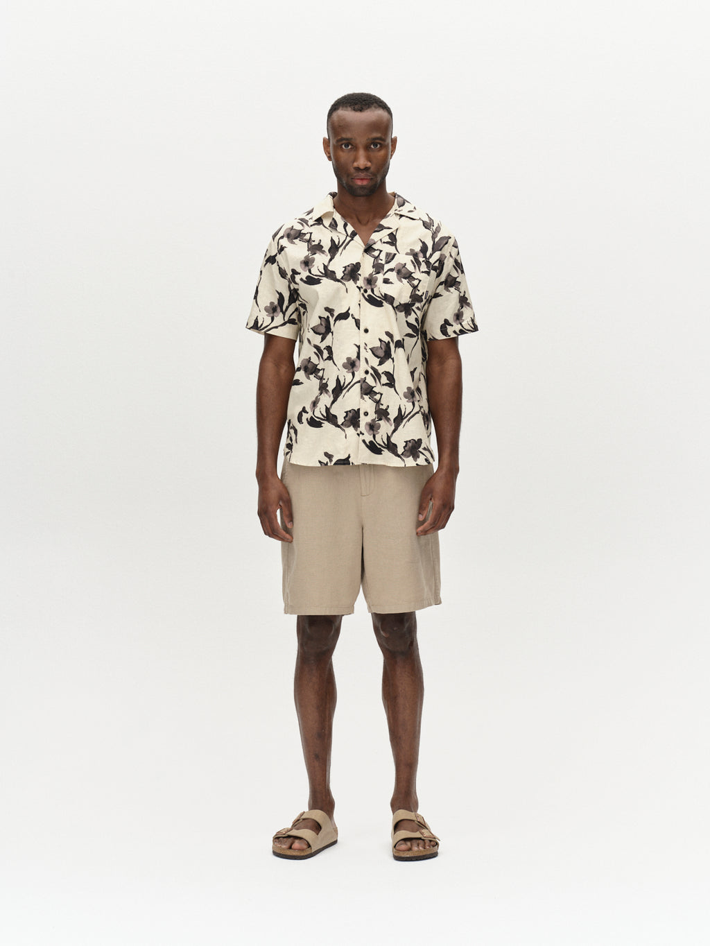 A person stands against a plain background in a Gabba Alfa Lino Sand Plaza short-sleeved shirt with dark shapes on a light base, premium shorts, and brown sandals. They gaze at the camera with a neutral expression, exuding an air of superior comfort.